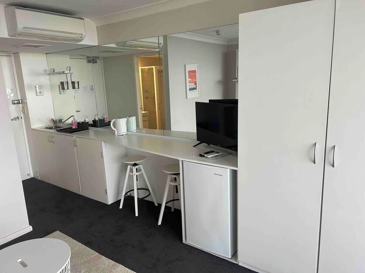 A compact hotel room with a kitchenette, a small bar area, and a single white stool.