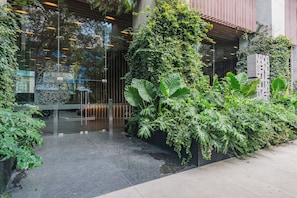 A modern building entrance with glass doors, a metal gate, and lush greenery including large leaves and vines.