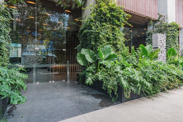 A modern building entrance with glass doors, a metal gate, and lush greenery including large leaves and vines.