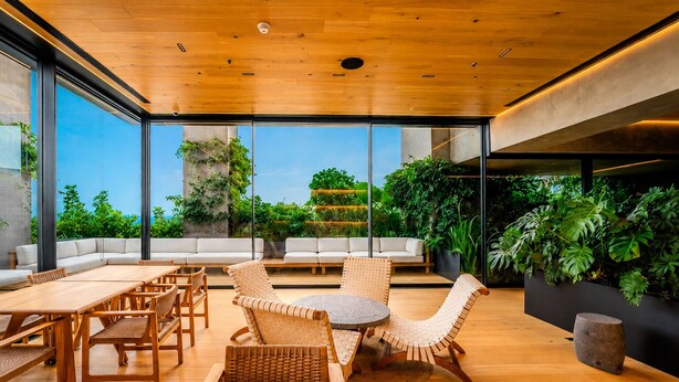 A modern outdoor lounge area with wooden furniture, large windows, and lush greenery. - The R Line I Sky Residences & Pool (Ciudad de México)