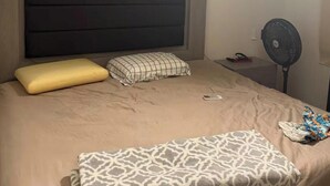 A bedroom with a bed, a bedside table, a fan, and a wall-mounted cross.