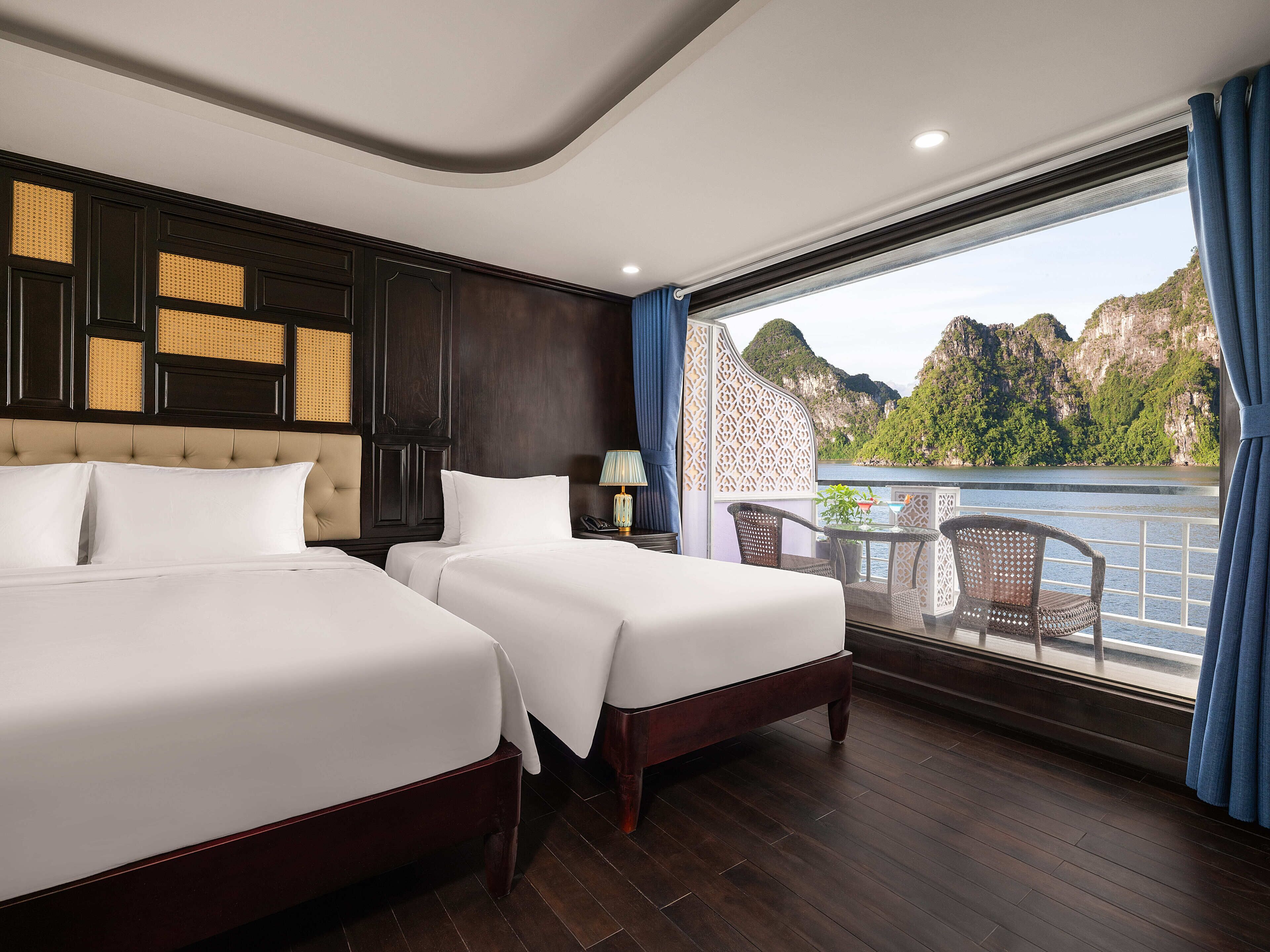 A hotel room with two beds, a balcony with a view of the water and mountains, and a wooden floor.