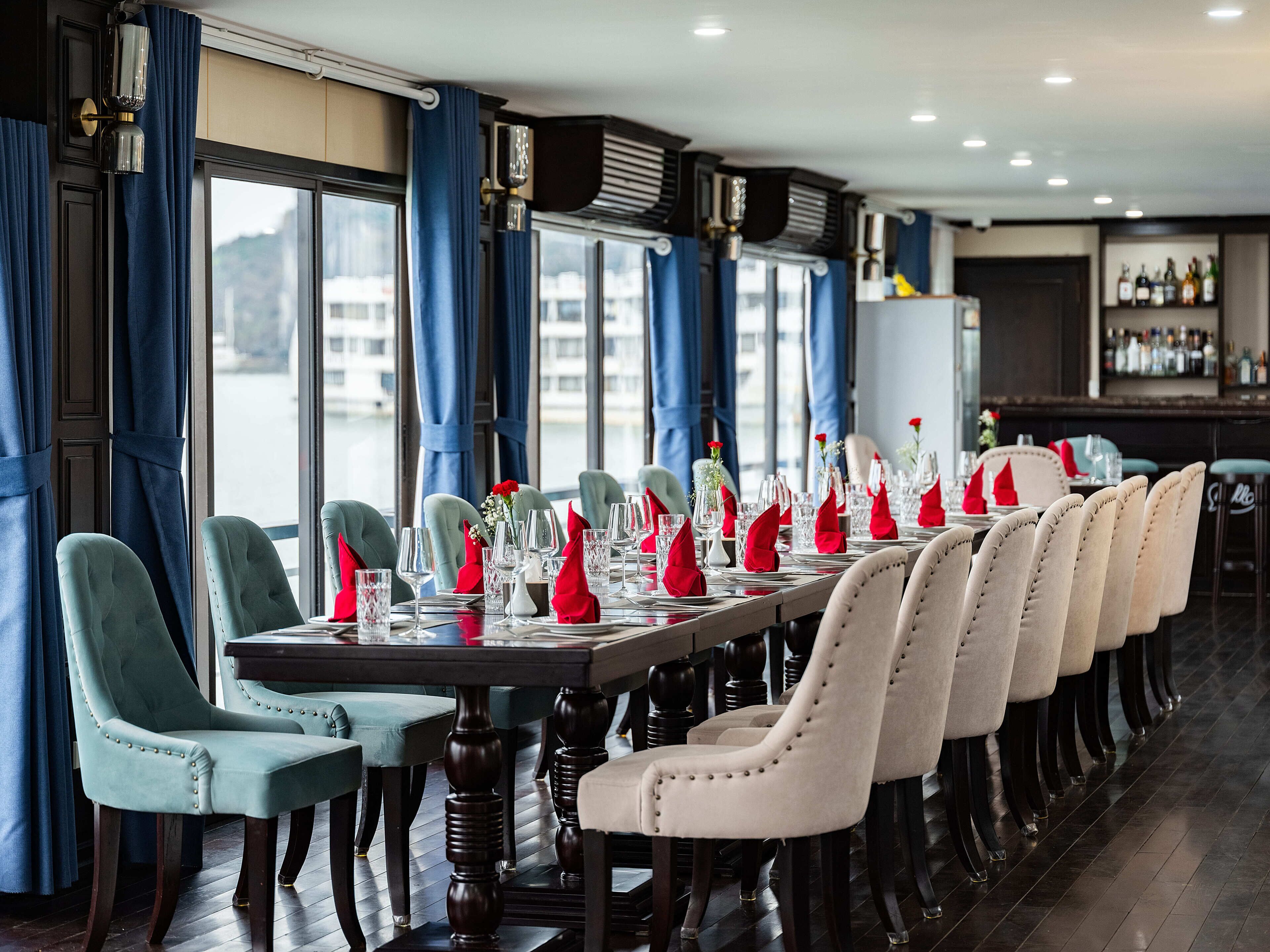 En restaurant med et langt spisebord dækket til et måltid, stole og et barområde med flasker.