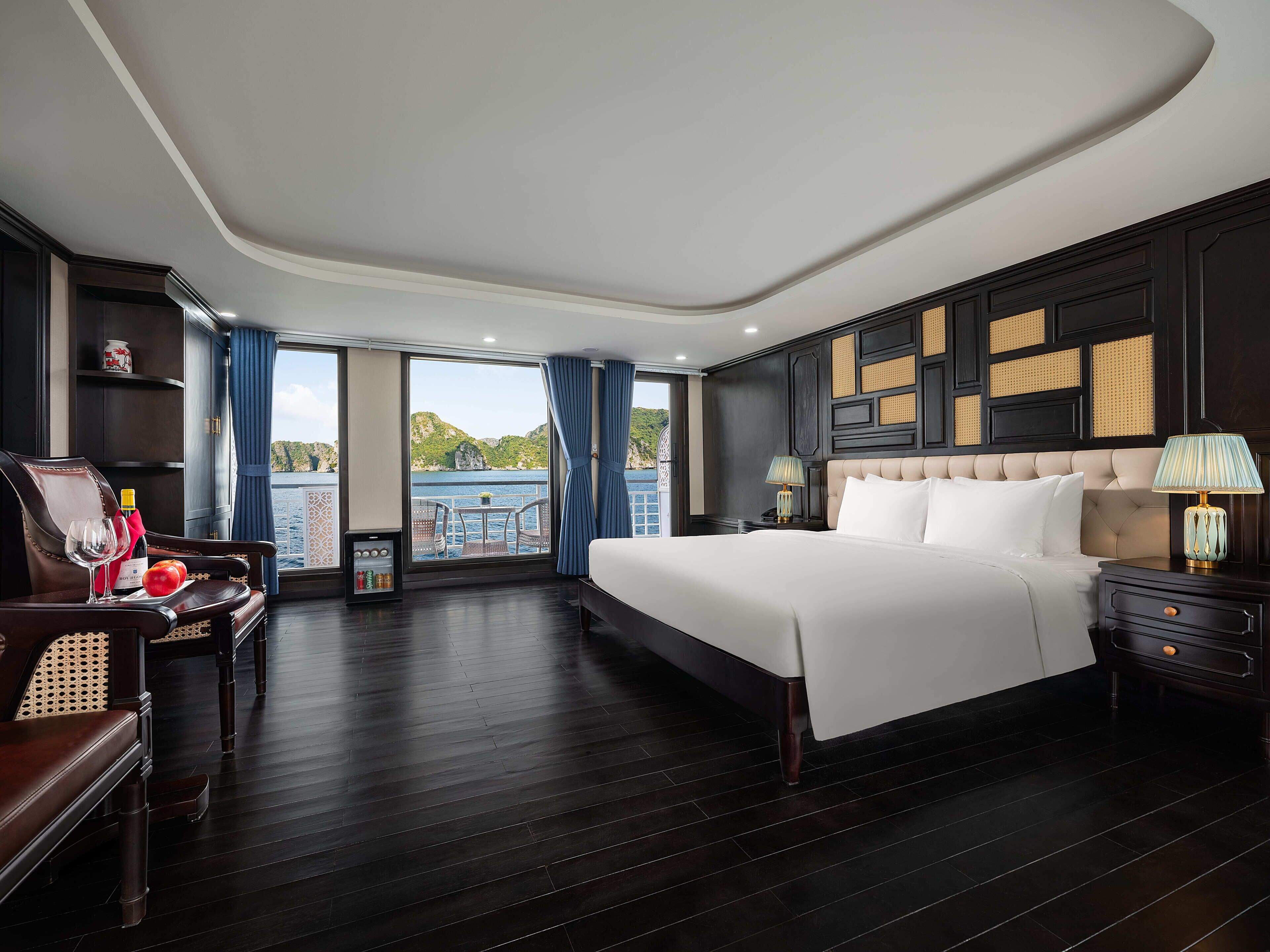 Habitación de hotel con una cama grande, zona de estar con mesa y sillas, mesita de noche con lámpara y vista al mar.