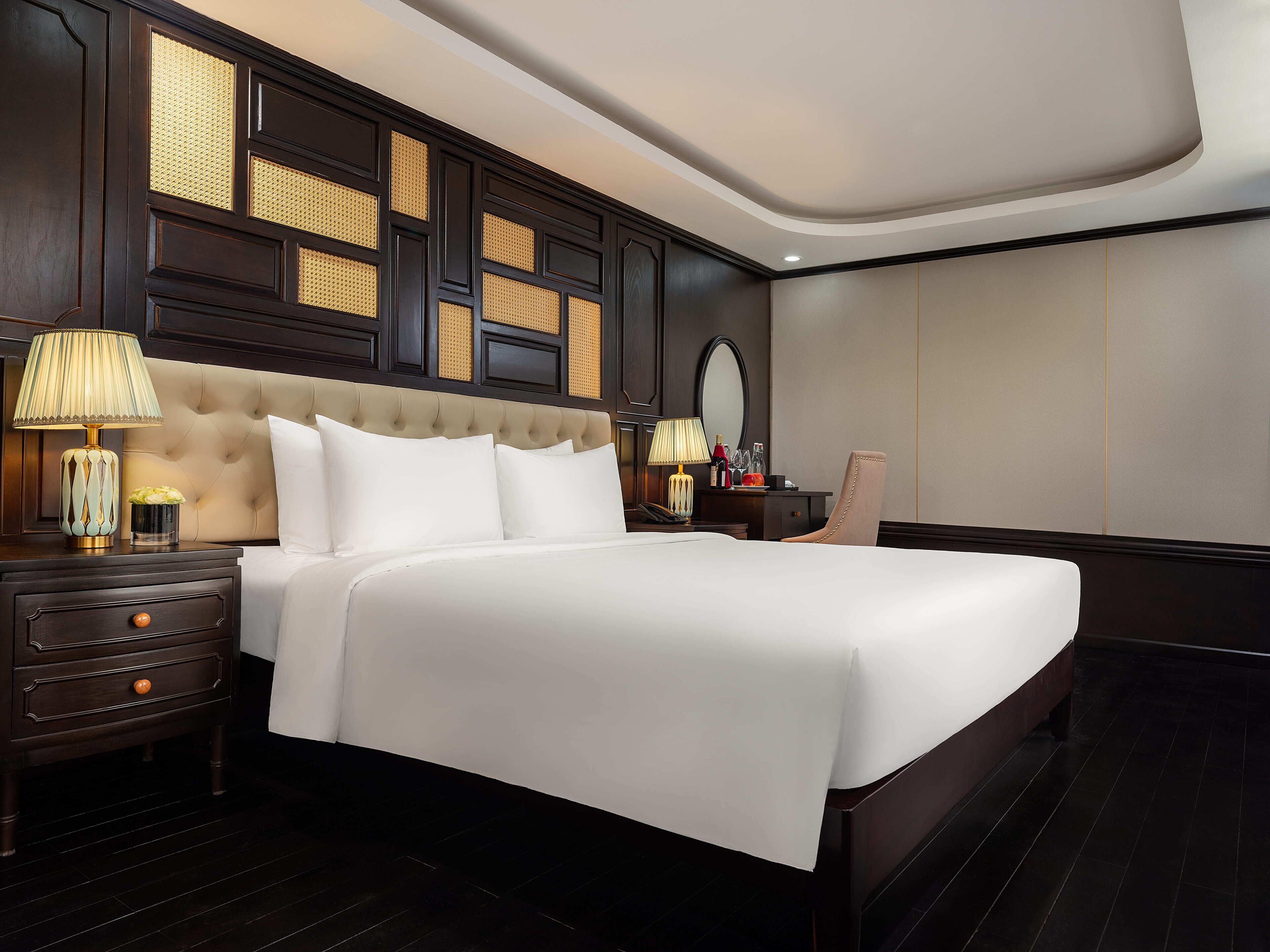 A hotel room with a large bed, dark wood furniture, a nightstand with a lamp, and a wall with a decorative panel.