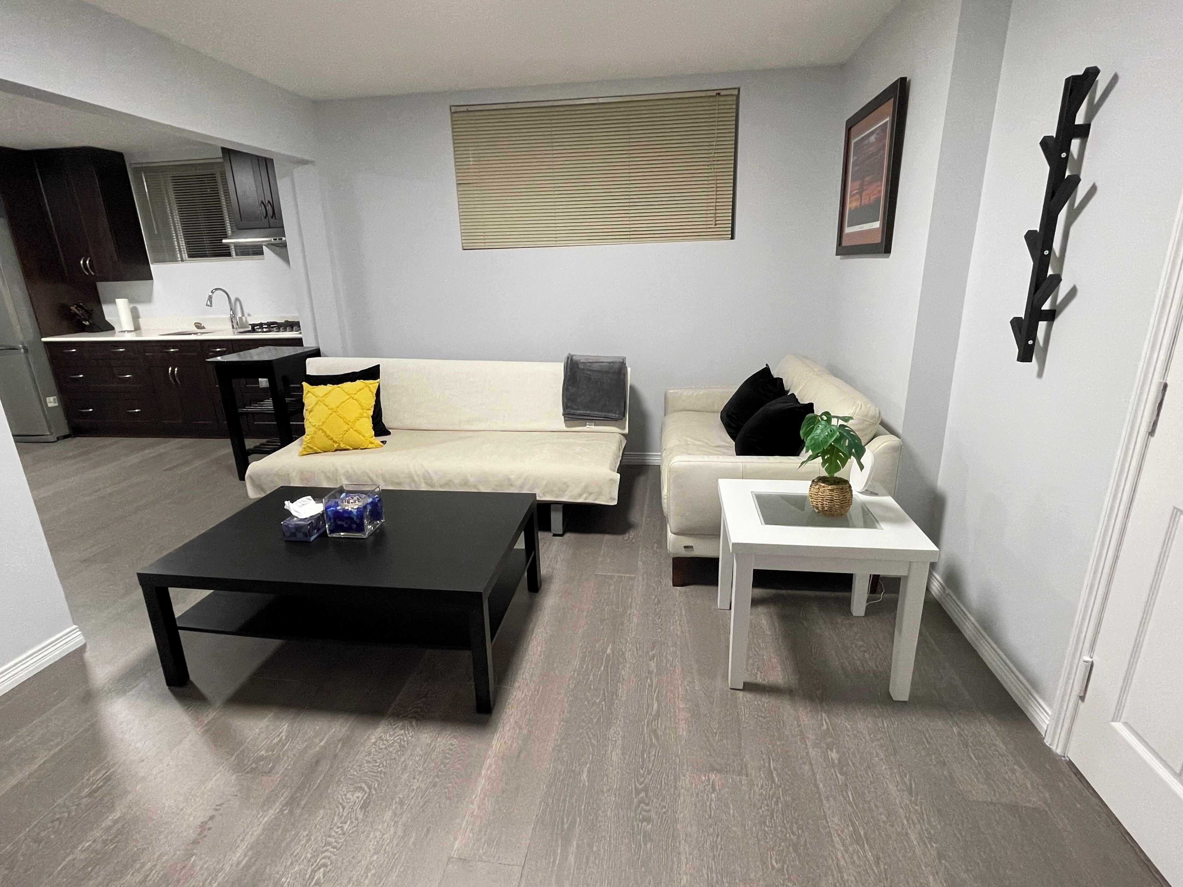 A living room with a sofa, a coffee table, and a side table. There is a kitchen area with cabinets and a window.