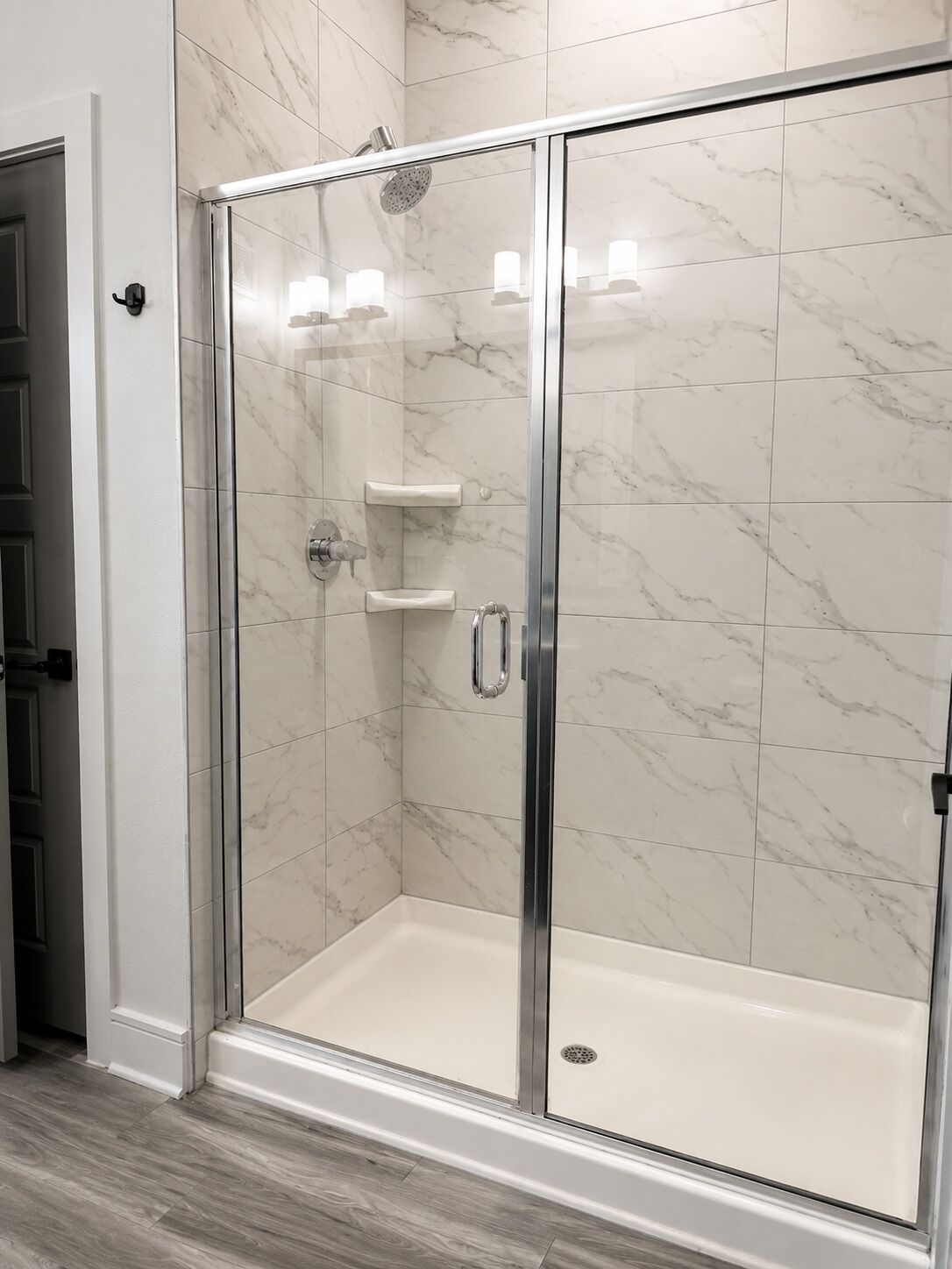 Une douche fermée par des parois de verre, avec un mur recouvert de carreaux de marbre blanc, une étagère intégrée et une porte dotée d’une poignée.