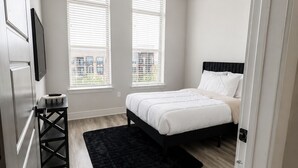 Een slaapkamer met een bed, een plafondventilator, twee ramen, een zwart vloerkleed en een televisie aan de muur.