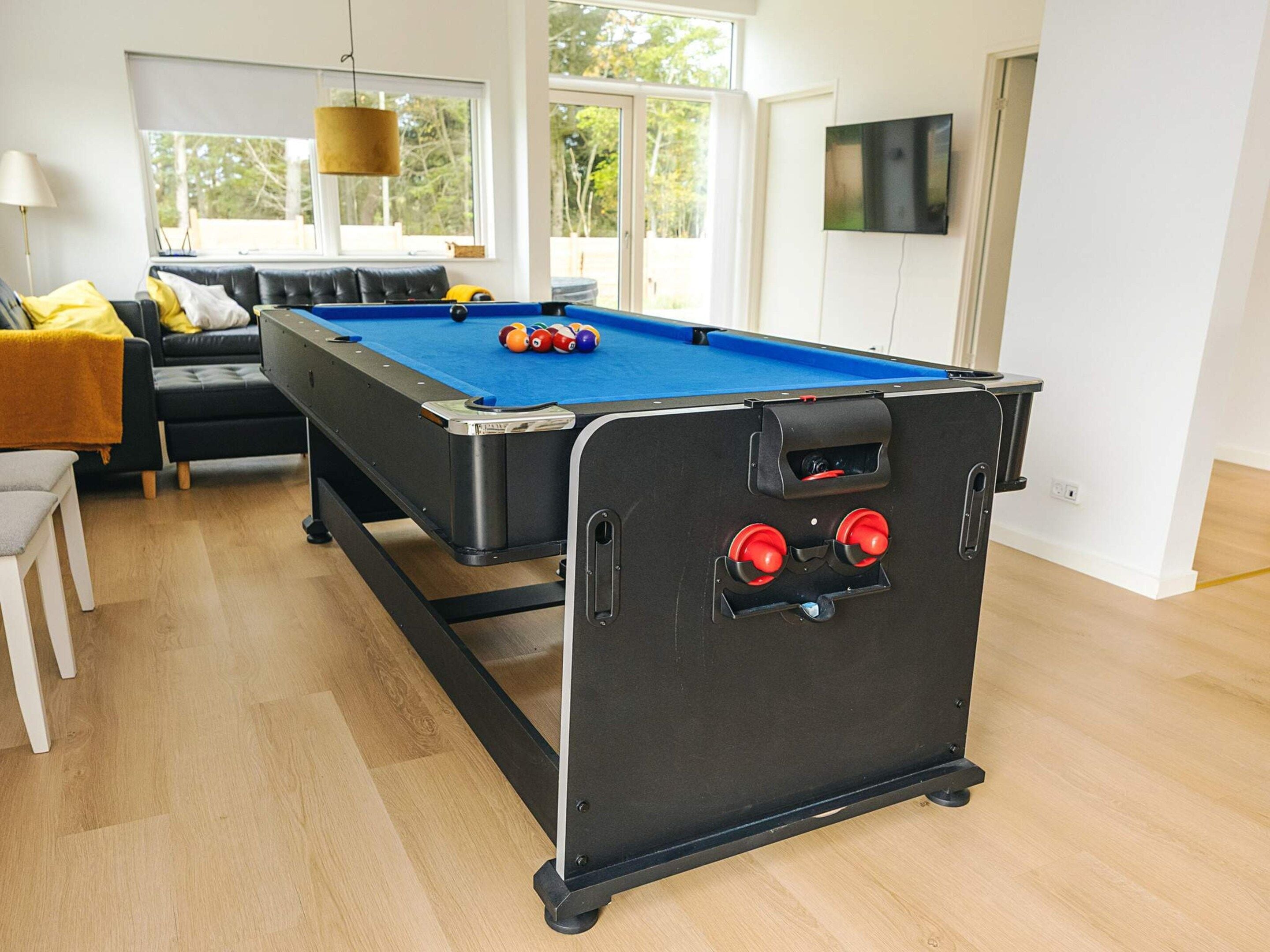 Een biljart tafel in een ruimte met een bank, een televisie en een raam met uitzicht op bomen.