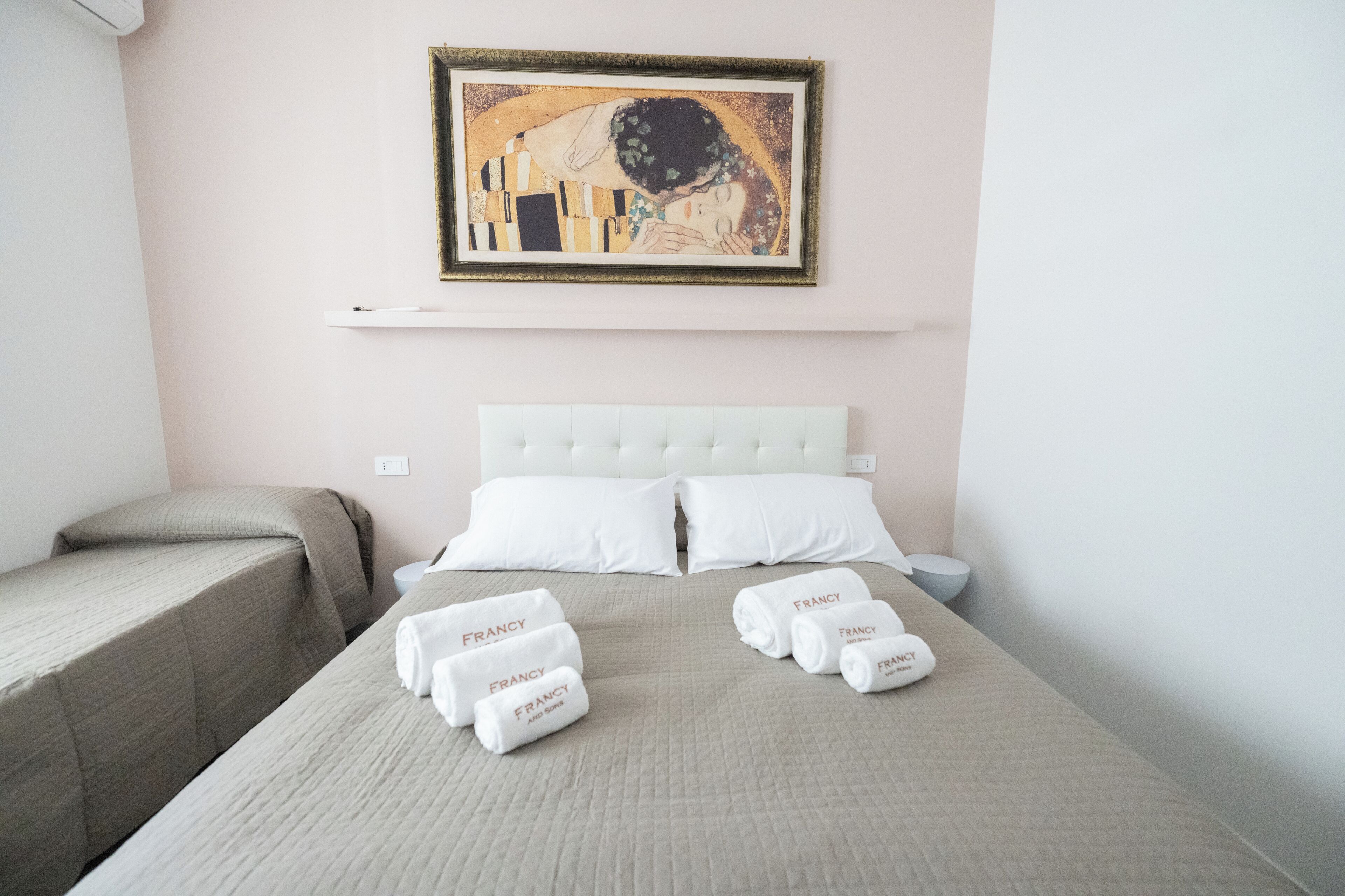 Habitación de hotel con cama, dos almohadas, cabecera, mesita de noche, un cuadro enmarcado en la pared y toallas con el nombre 'FRANCY' sobre la cama.