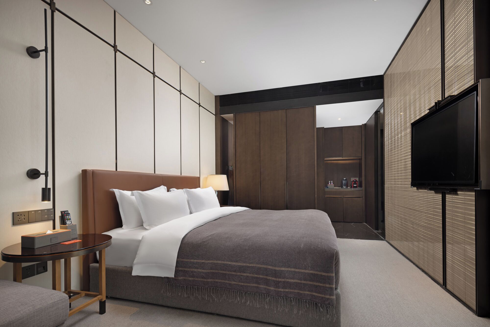 A modern hotel room with a large bed, bedside table, lamp, and a flat-screen TV mounted on the wall.