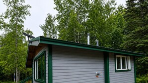 Eine kleine, eingeschossige Holzhütte mit einem grünen Dach und weissen Verkleidungen, einer Veranda mit Holzgeländer und einem Schornstein.