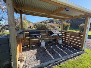 Zona de barbacoa al aire libre con una parrilla, un tanque de gas y un letrero que indica limpiar las barbacoas después de usarlas.