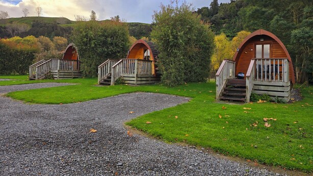 Three wooden cabins with wooden railings and steps, set in a grassy area with trees and hills in the background.