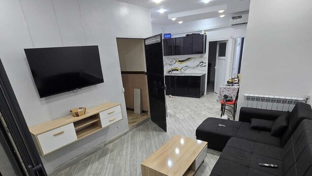 Ein modernes Wohnzimmer mit einem Flachbildfernseher, einem schwarzen Sofa, einem Holztisch und einer Küche im Hintergrund.