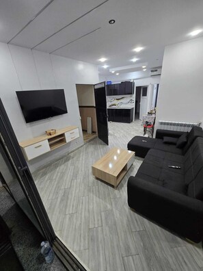 Una sala de estar moderna con una televisión de pantalla plana, un sofá negro, una mesa de centro de madera y una cocina al fondo.