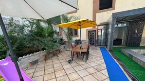Eine Terrasse im Freien mit Tisch und Stühlen, einem gelben Sonnenschirm, einer violetten Rutsche und einem blauen Teppich.
