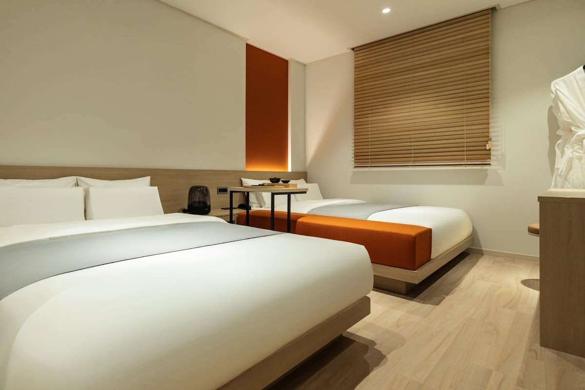 Ein Hotelzimmer mit zwei Betten, einem Schreibtisch, einem Sessel, einem Nachttisch, einem Fenster mit Jalousien und einer Statue.