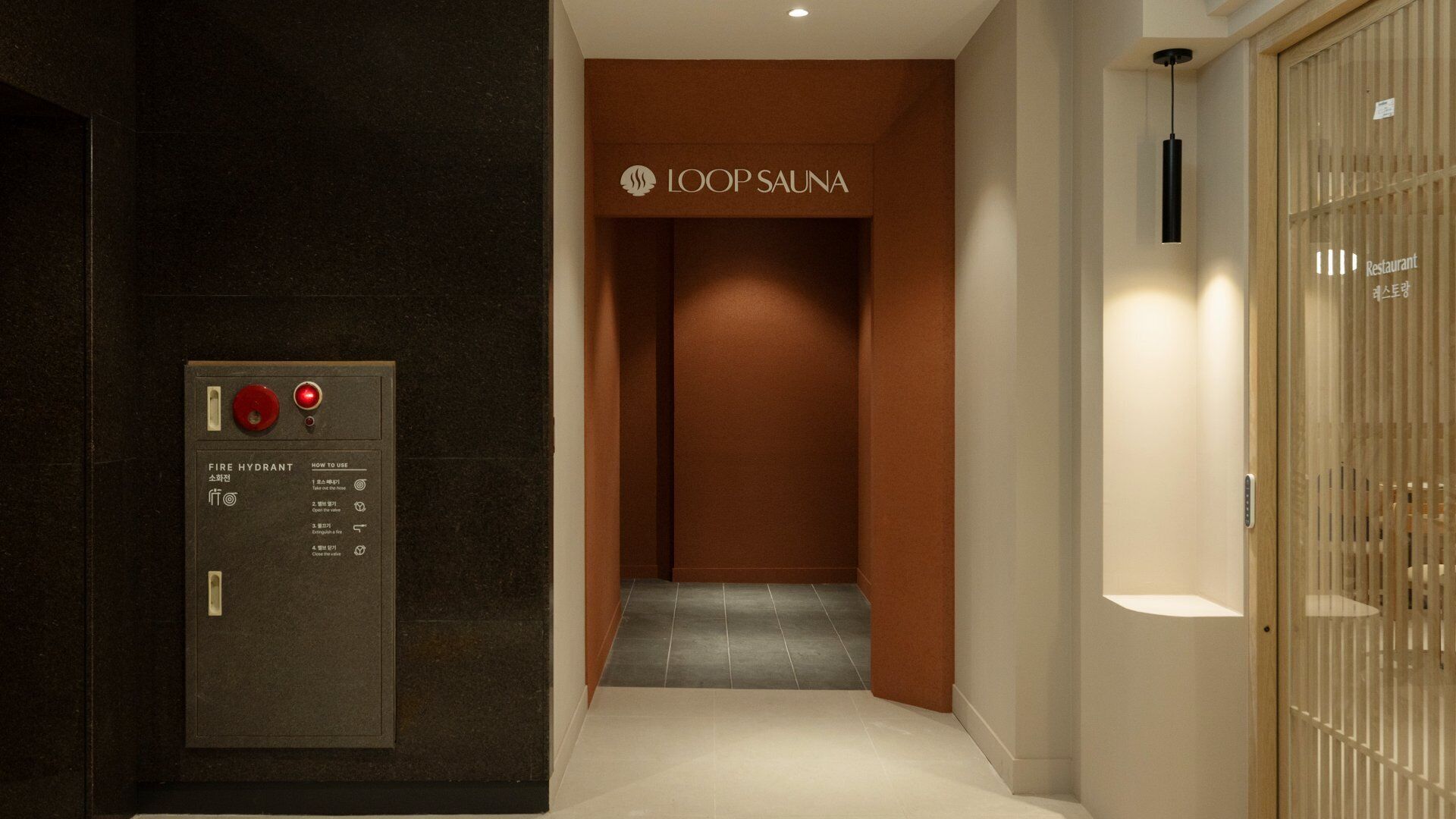 A hallway with a fire hydrant control panel, a sign for LOOP SAUNA, and a wooden door labeled 'Restaurant'.