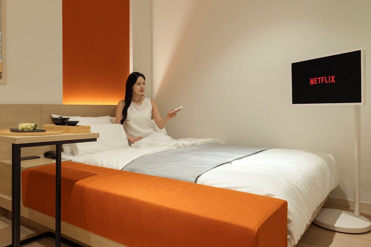 Ein modernes Hotelzimmer mit einem Bett, einem Nachttisch mit Fernbedienung und einem am Wand montierten Fernseher, der 'NETFLIX' anzeigt.