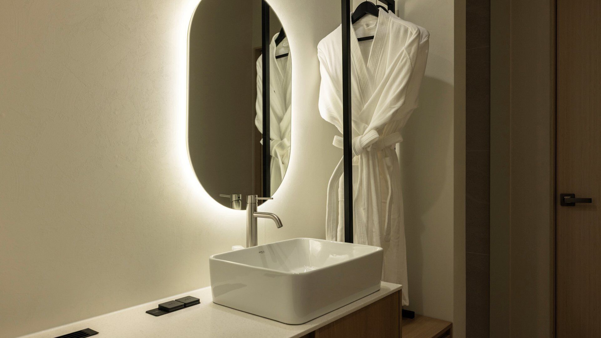 Een badkamer met een rechthoekig wastafelmeubel, een spiegel met een ovale lijst, en een badjas hangend aan een rek.