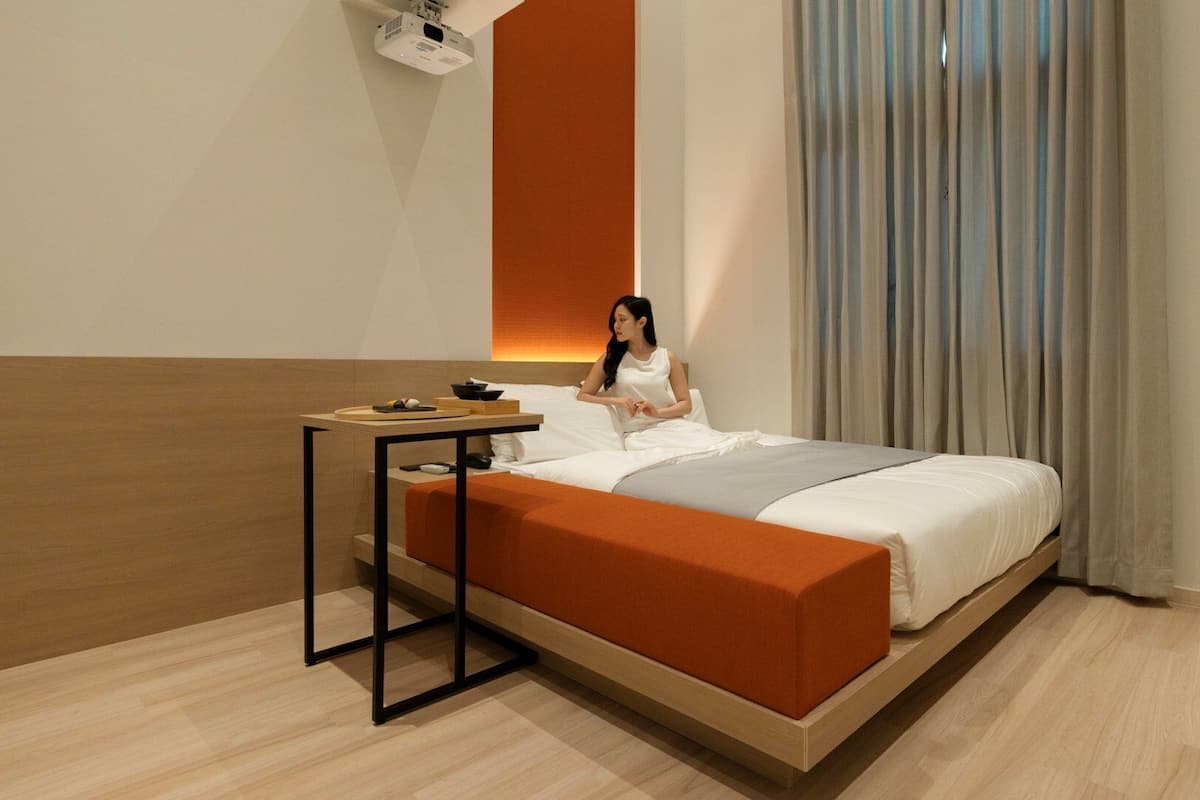 Ein modernes Hotelzimmer mit einem Bett, einem Nachttisch, einem an der Wand montierten Projektor und einem großen Fenster mit Vorhängen.
