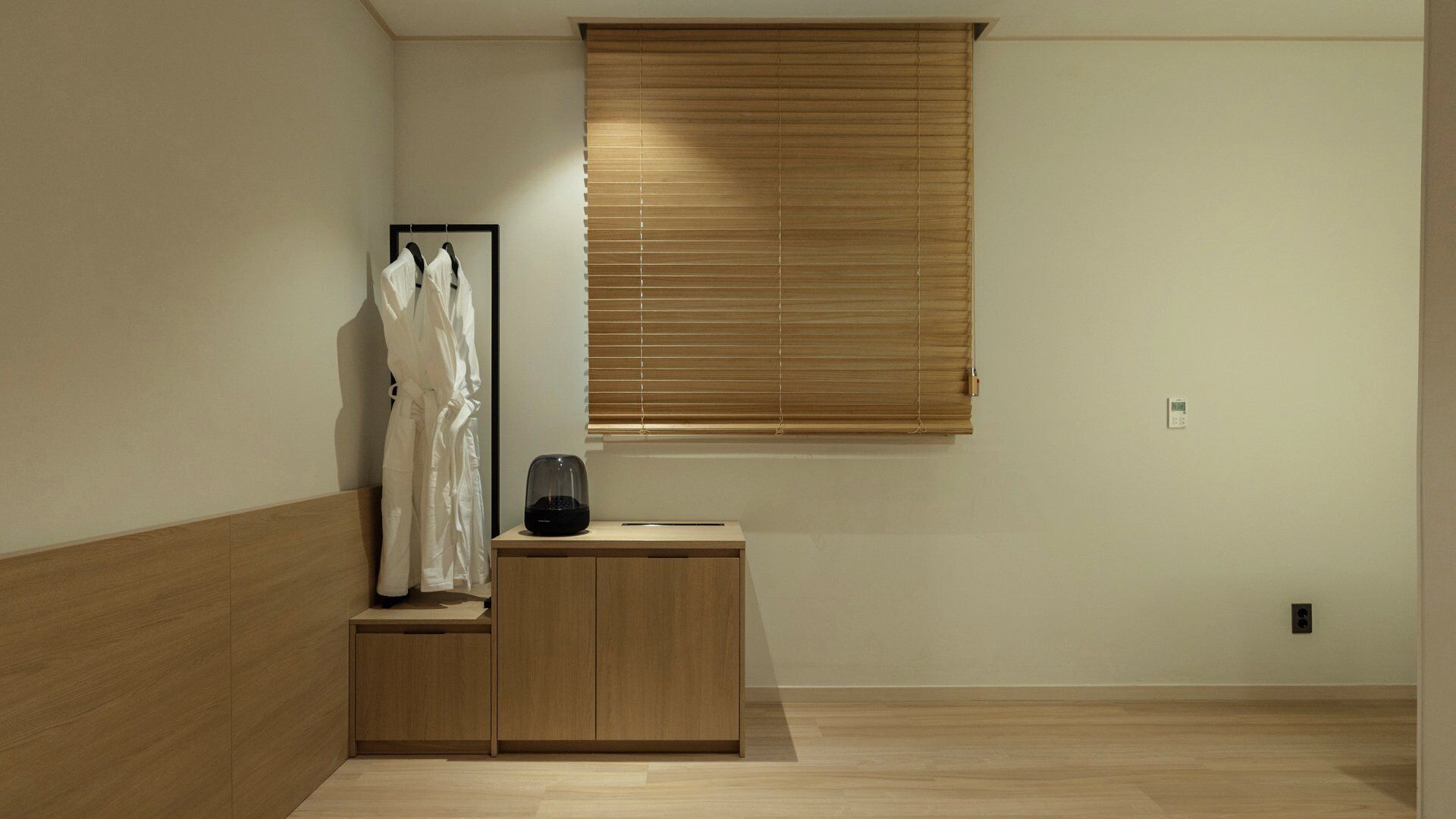 Una habitación con piso de madera, un armario de madera, un parlante negro y una ventana con persianas.