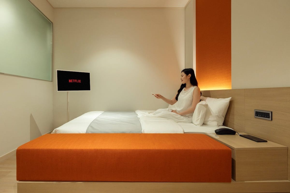 Ein modernes Hotelzimmer mit einem Bett, einem Nachttisch mit Fernbedienung und einem an der Wand befestigten Fernseher, der 'NETFLIX' anzeigt.