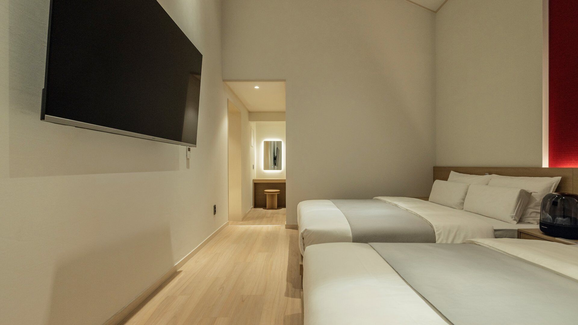 A hotel room with two beds, a TV, a bathroom with a mirror, and a red accent wall.