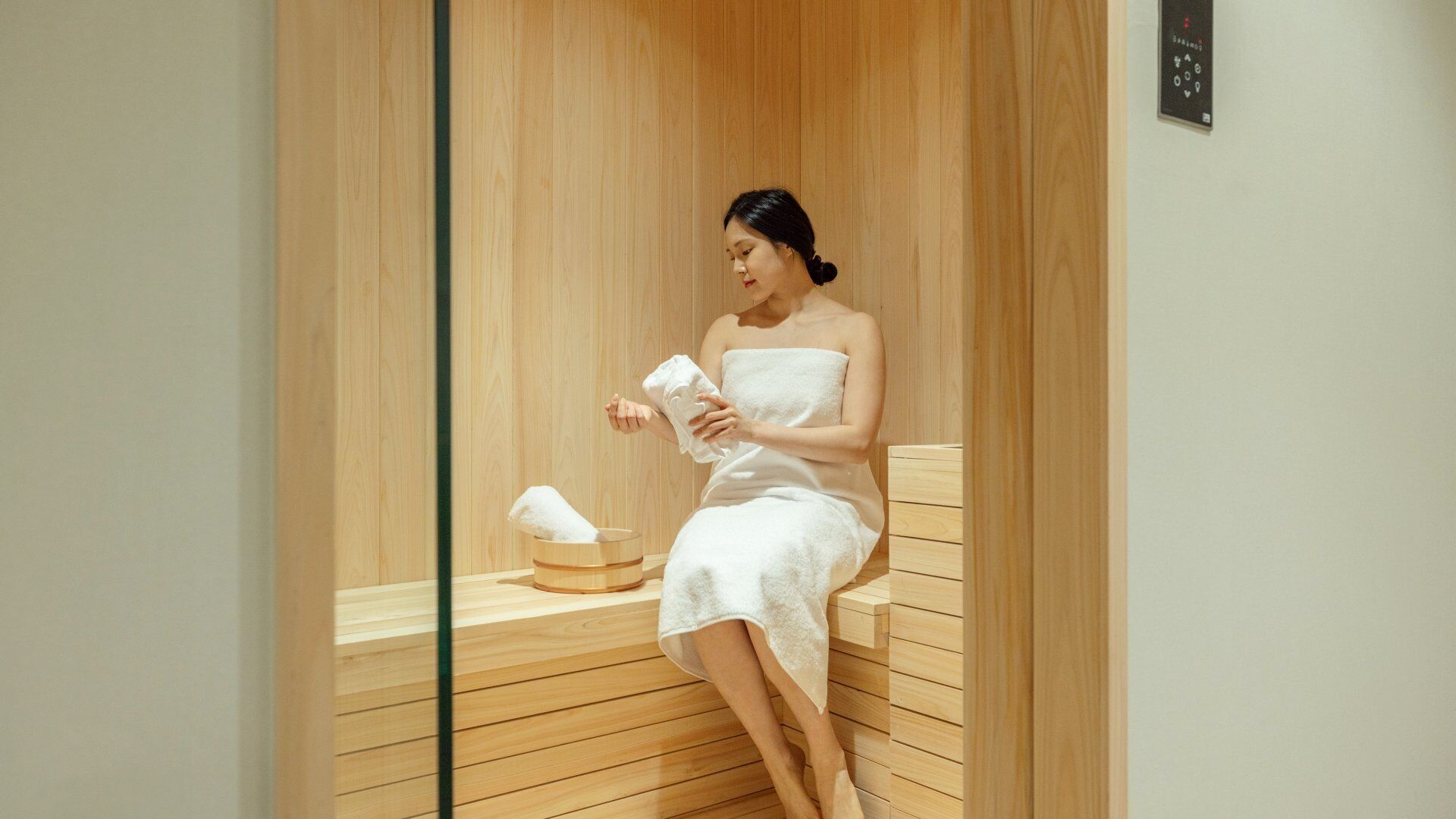 A person in a white towel sitting in a wooden sauna with a towel on the bench and a control panel on the wall.