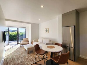 A modern kitchen with a large refrigerator, a dining table with chairs, a sofa, and a TV mounted on the wall. - Serenity Sands Apartment - wheelchair friendly, at reserve, close to beach (Hahei)