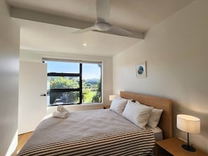 A bedroom with a bed, a nightstand, a lamp, a ceiling fan, a window with a view, and a door. - Serenity Sands Apartment - wheelchair friendly, at reserve, close to beach (Hahei)