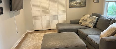 A living room with a grey sofa, an ottoman, a white closet, and a wall-mounted TV.