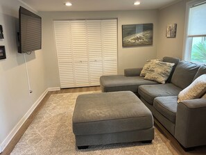 Un salon avec un canapé gris, un pouf, une armoire blanche et un téléviseur fixé au mur.