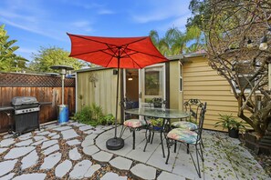 Eine Terrasse mit Glastisch und -stühlen, einem roten Sonnenschirm, einem Grill und einem gepflasterten Weg.