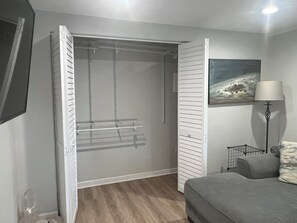 A bedroom with a grey sofa, a bedside table with a lamp, a hanging coat rack, and a painting on the wall.