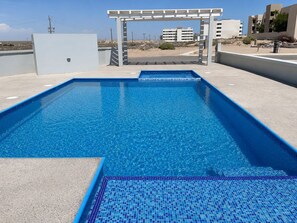 Ein rechteckiger Pool mit einer mosaikbesetzten Kante, einem Pergolen-Anbau und Blick auf Gebäude im Hintergrund.