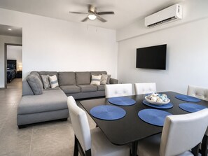 Ein modernes Wohnzimmer mit einem grauen Ecksofa, einem Esstisch mit blauen Tischsets, weißen Stühlen, einem Deckenventilator und einem wandmontierten Flachbildfernseher.