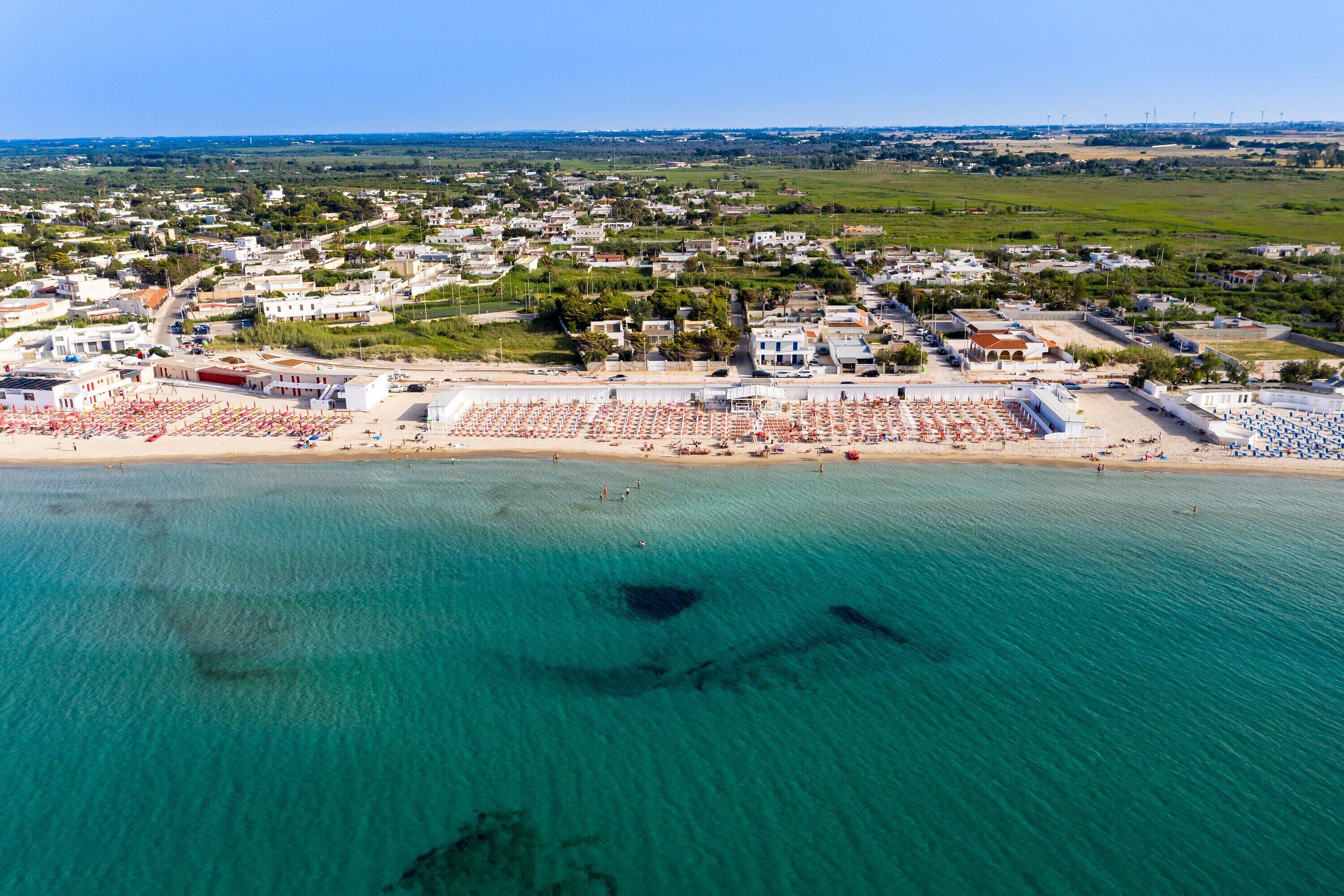 Vue aérienne d’une ville côtière avec une plage, des bâtiments et une eau d’un bleu limpide.