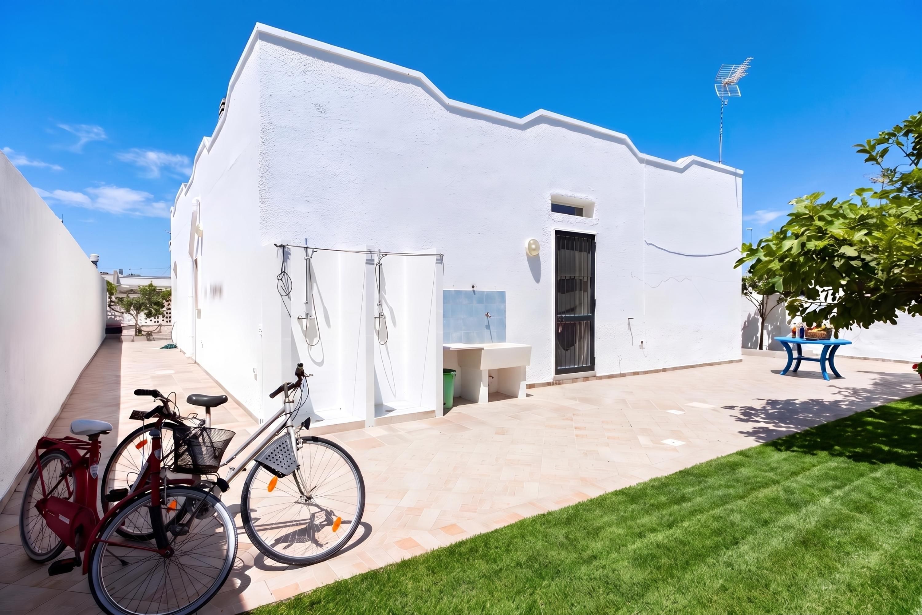 A white building with a flat roof, a small window, and a door. There is a bicycle parked in front and a table with a plant on the right.