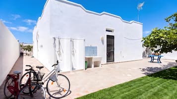 A white building with a flat roof, a small window, and a door. There is a bicycle parked in front and a table with a plant on the right.