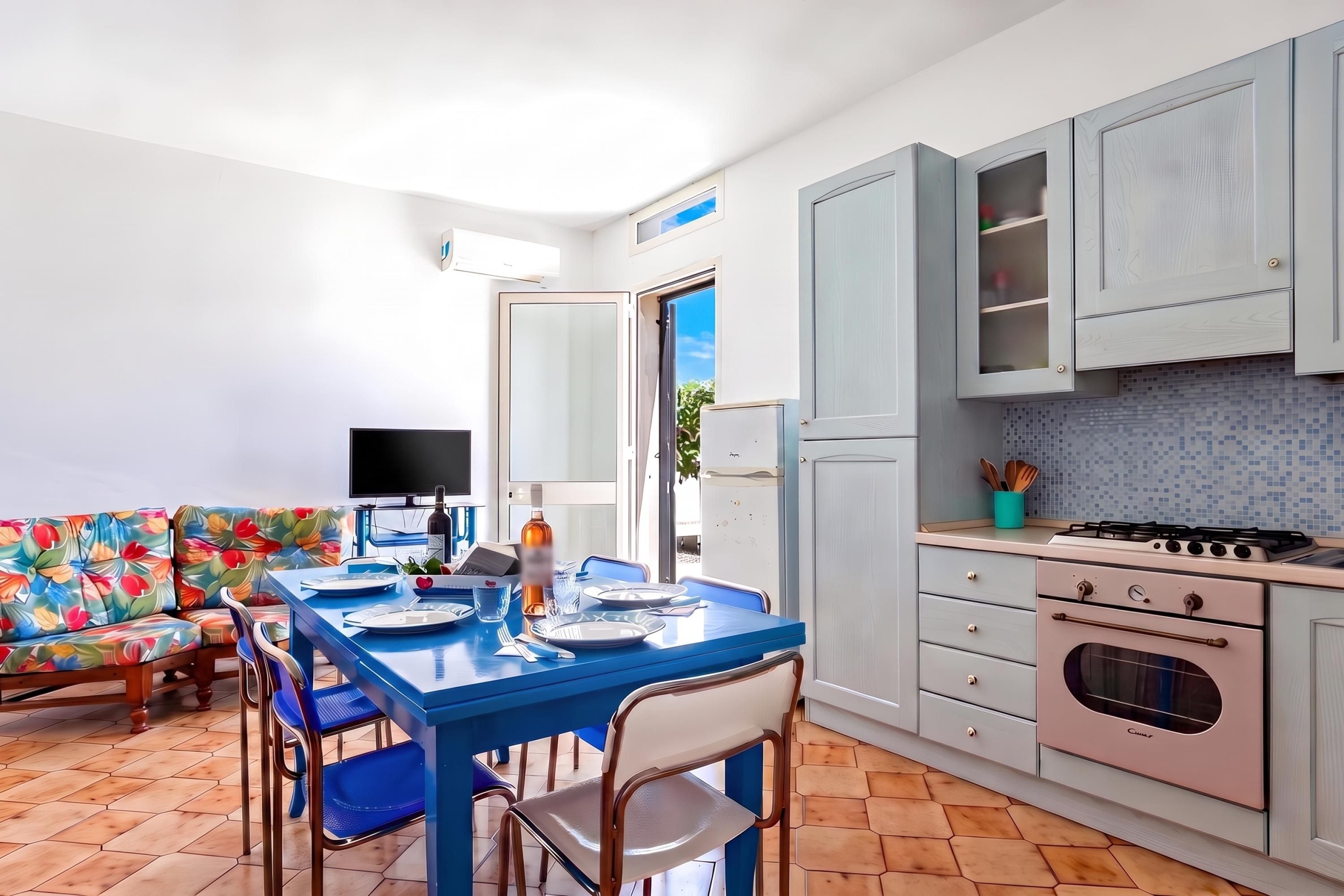 A modern kitchen with a dining area, featuring a blue table set for two, a sofa with a colorful floral pattern, and a large window.