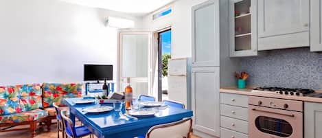 Una cocina moderna con comedor, que presenta una mesa azul dispuesta para dos personas, un sofá con estampado floral colorido y un ventanal grande.