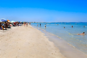 Ein Strand mit badenden Personen und Liegestühlen, Sonnenschirmen und einem klaren Himmel.
