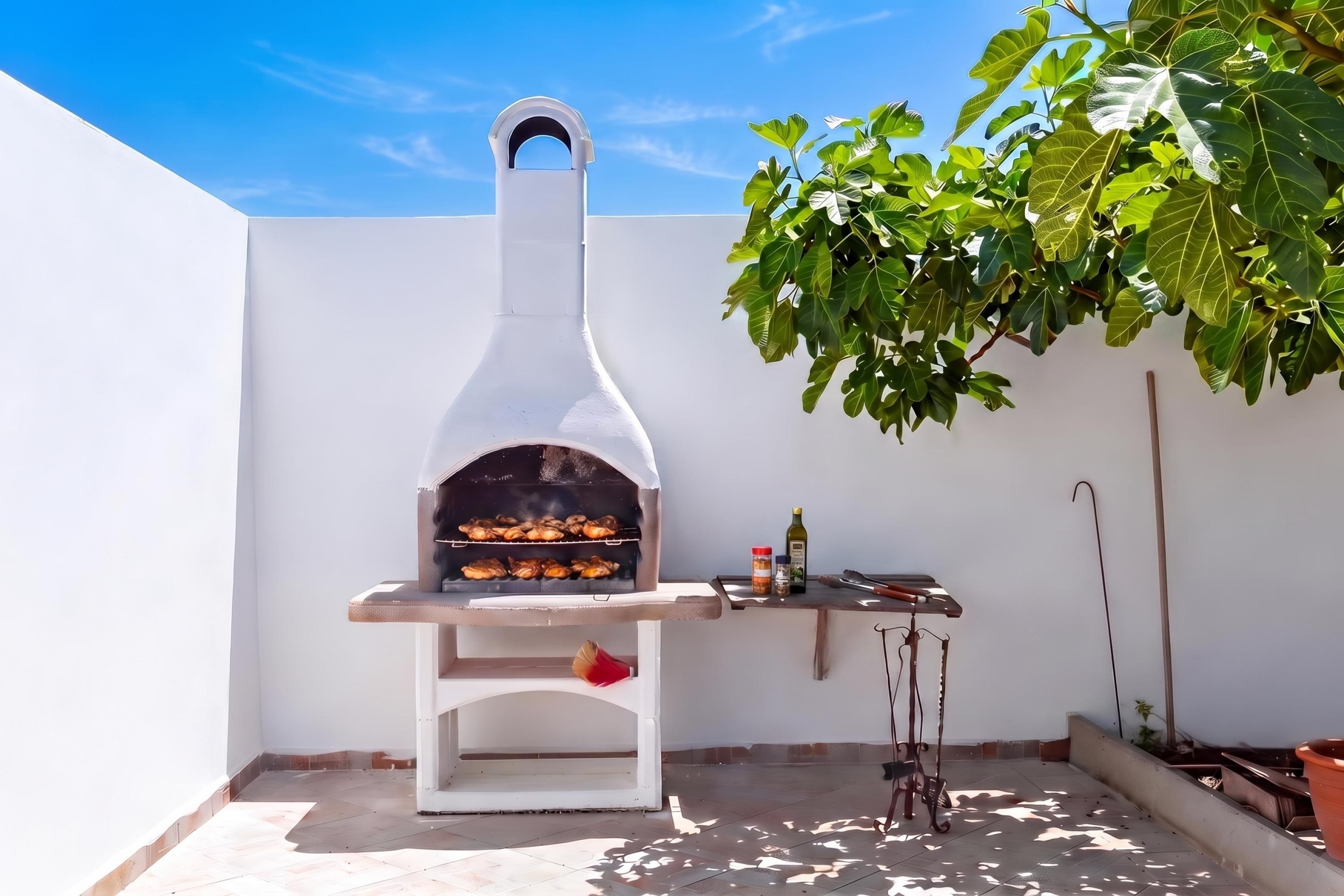 Forno de pizza branco ao ar livre com chaminé, mesa com garrafa de azeite e pequeno recipiente de especiarias.