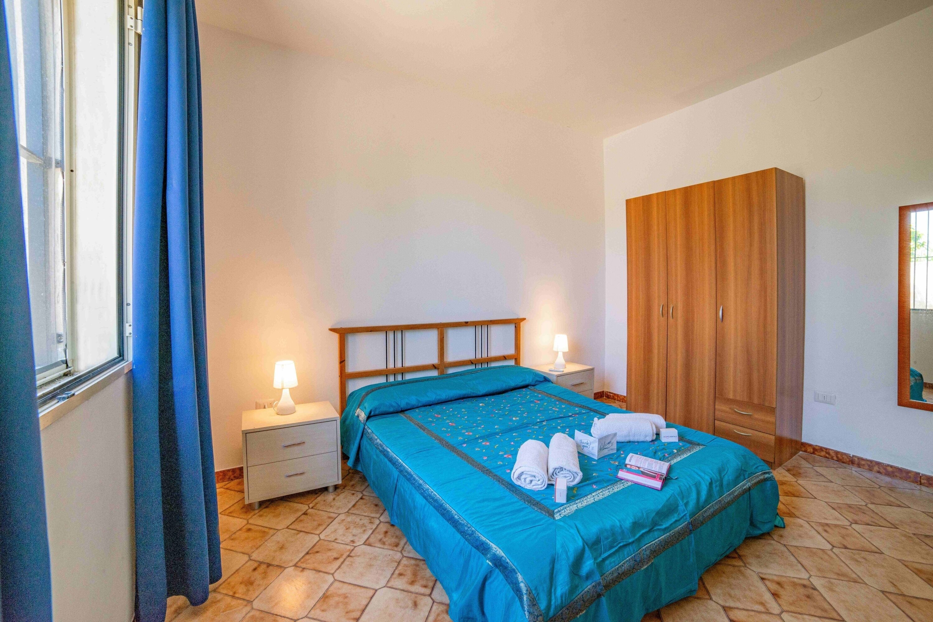 Quarto com cama, mesa de cabeceira, guarda-roupa e janela com cortinas.