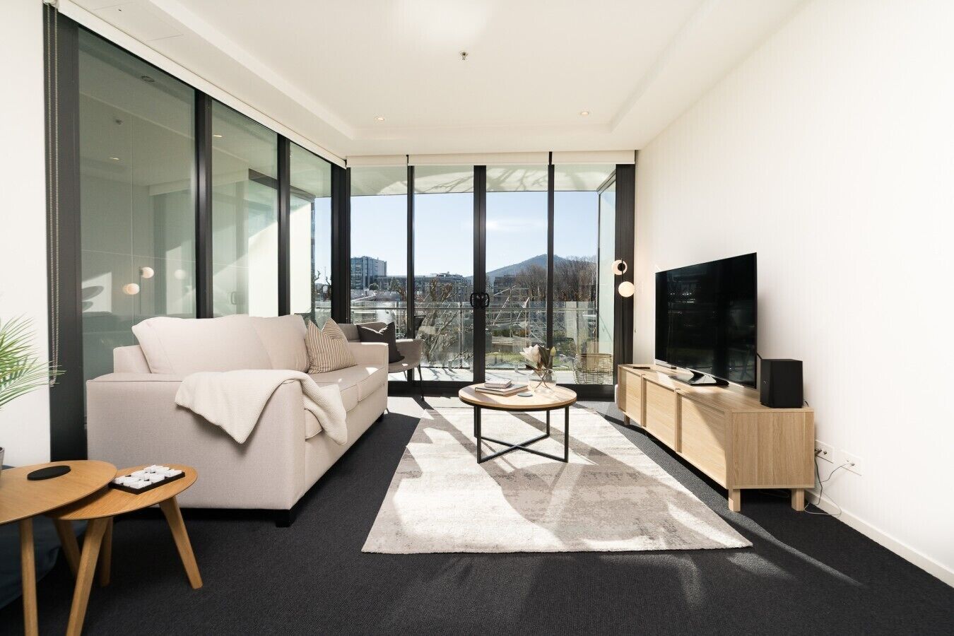 A modern living room with a sofa, coffee table, TV, and a view through large windows.