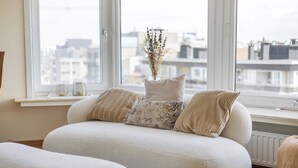 Un soggiorno moderno con un divano, un vaso con fiori secchi e vista sugli edifici attraverso ampie finestre.