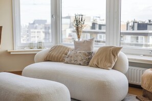 Ein modernes Wohnzimmer mit einer Couch, einer Vase mit getrockneten Blumen und Blick auf Gebäude durch große Fenster.
