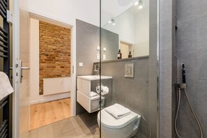 Une salle de bain moderne avec une douche à parois de verre, des toilettes blanches, un lavabo avec un miroir et un mur en briques apparentes.